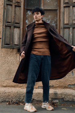 Kahverengi ceketli, beyaz tişörtlü, kot pantolonlu ve beyaz ayakkabılı bir adam bir binanın önünde duruyor..