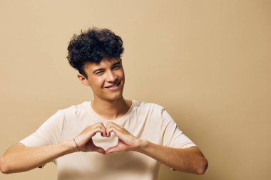 A young male figure stands against a soft beige background, forming a heart shape with his hands His cheerful expression and relaxed posture create a warm atmosphere, perfect for themes of love clipart