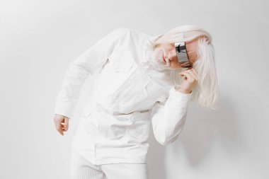 Tamamen beyaz bir takım elbise ve güneş gözlüklü şık bir model, modernliği temsil eden minimalist beyaz bir arka plana karşı fütürist ve çağdaş modanın bir karışımını sergiliyor.