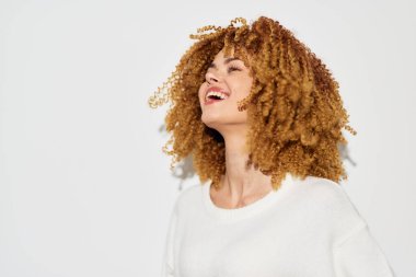 Kıvırcık sarı saçlı neşeli bir kadın sade beyaz bir arka plana gülüyor, mutluluk ve kendine güven duygusu yayıyor, kaygısız bir ruhu somutlaştırıyor ve fotoğrafçılıkta canlandırıcı bir ruh hali sergiliyor.