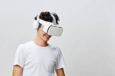 Kıvırcık saçlı genç adam sanal gerçeklikten zevk alıyor, beyaz bir VR kulaklık ve kulaklık takıyor, hafif bir arka plana karşı modern teknolojiyi ve büyüleyici deneyimleri temsil ediyor.
