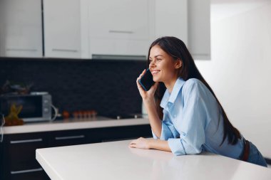 Ev ortamında mutfak masasında oturan bir kadın cep telefonuyla konuşuyor.