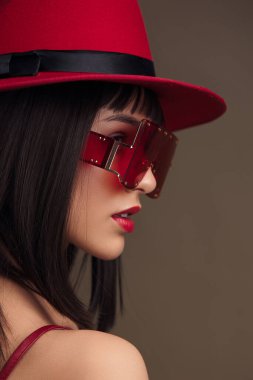 Şık kırmızı şapkalı ve güneş gözlüklü bir kadın koyu kahverengi bir arkaplanda güven içinde poz veriyor, çarpıcı ve zarif bir moda imajı yaratıyor.