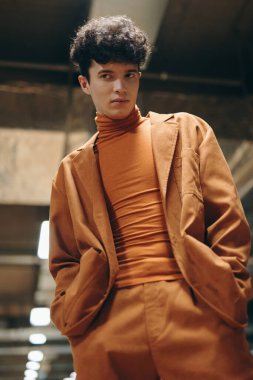 Şık, balıkçı yaka ve büyük beden ceket giyen genç bir adam havalı ve modern bir ortamda rahatça poz veriyor.