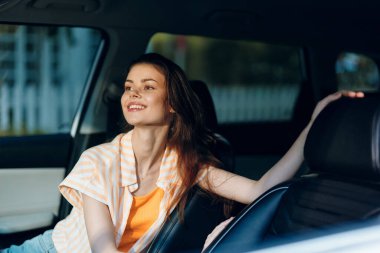 Neşeli genç bir kadın arabada oturuyor, pencereden dışarı bakarken mutluluk saçıyor uzun saçları var ve sıradan çizgili bir bluz ve turuncu gömlek giyiyor.