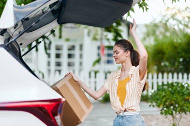 Uzun saçlı, çizgili gömlekli bir kadın, bir arabanın bagajına karton bir kutu yerleştiriyor. Güneşli bir açık hava ortamında geçiyor. Teslimat, lojistik ve günlük işleri yansıtıyor.