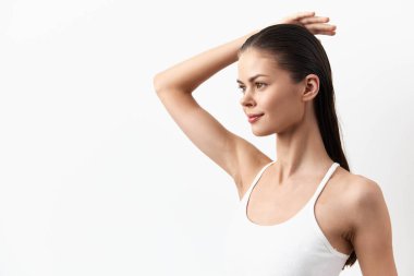 Beyaz atletli genç bir kadın beyaz arka planda zarif bir poz veriyor. Uzun saçları düz ve pürüzsüz. Kolunu zarifçe kaldırırken düşünceli bir yüz ifadesi takınıyor.