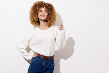 Kıvırcık saçlı, rahat beyaz bir süveter ve yüksek bel kot pantolon giyen, tarafsız, hafif bir arka plana karşı duran, sıradan ve şık bir modayı temsil eden mutlu genç bir kadın.
