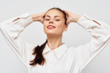 Bu resim beyaz gömlekli, kendine güvenen genç bir kadını, elleri başının arkasında poz verirken büyüleyici bir şekilde gülümserken gösteriyor. Açık gri arka plan onun zarafetini arttırıyor, bu da onu uygun kılıyor.