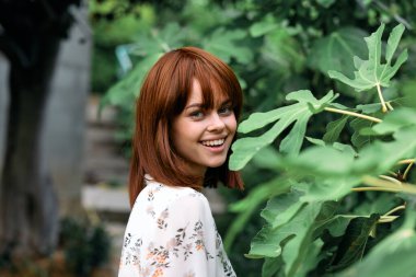 Canlı kızıl saçlı kadın yemyeşil bir yerde duruyor, doğal ortamında kameraya gülümsüyor.