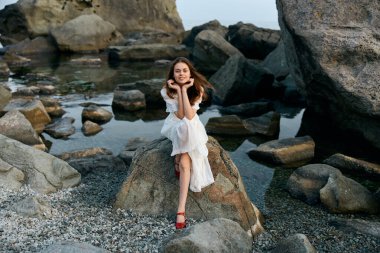 Beyaz elbiseli sakin bir kadın okyanus kayalığında dizlerinin üstünde oturmuş dalgaları seyrediyor.