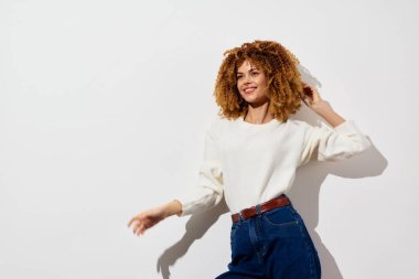 Şehvetli kıvırcık saçlı neşeli genç bir kadın minimalist beyaz bir arka fonda duruyor rahat beyaz bir kazak ve mavi yüksek bel kot pantolon giyiyor kendinden emin duruşu mutluluğu yayıyor