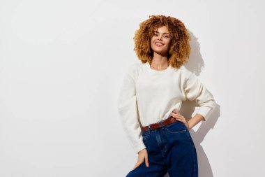 Kıvırcık saçlı şık bir bayan model, rahat bir süveter ve yüksek bel kotu giymiş, düz beyaz bir duvara karşı güven içinde poz veriyor, neşeli bir hava yayıyor.