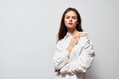 Genç bir kadın, kollarını kavuşturmuş, beyaz bir gömlekle arka planda durarak özgüvenle duruyor. İfadesi düşünceli ve kendinden emin, modern zarafeti somutlaştıran ve dengeli.