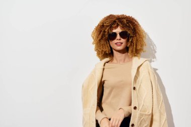 Havalı kıvırcık saçlı, bej bluzlu ve büyük beden hırkalı, güneş gözlüklü, kendine güvenen, sade beyaz arka planda, fas için ideal bir kadın.