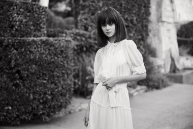 Beyaz elbiseli zarif bir kadın yemyeşil çalıların önünde duruyor.