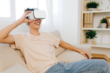 Sanal gerçeklik kulaklığı takan neşeli genç adam, evde engin bir deneyimin tadını çıkarıyor açık renkli oda yeşillik barındırıyor ve rahatlamış bir havayı koruyor, oyun ve yenilik için ideal