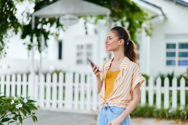 Gülümseyen genç bir kadın dışarıda güneşli bir günün tadını çıkarırken akıllı telefonunu kullanıyor. Çizgili bir gömlek ve günlük şort giyiyor. Etrafı yeşil ve beyaz çitlerle çevrili.