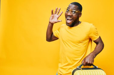 Gülümseyen genç bir adam parlak sarı bir arka planda bavul tutuyor, neşe ve heyecan saçıyor günlük sarı bir tişört giyiyor ve şık gözlükler takıyor, neşeli bir seyahat havası yakalıyor.