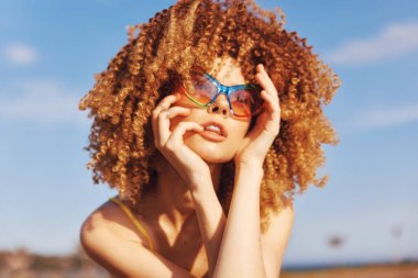 Parlak kıvırcık saçlı şık bir genç kadın, güneş gözlüğü takarken güneşin tadını çıkarıyor, kaygısız bir hava yaratıyor.