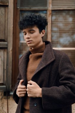 Kahverengi paltolu, kıvırcık saçlı ve boynu kaplumbağa gibi süveterli, ahşap kapılı şık bir genç adam pencerenin önünde duruyor..