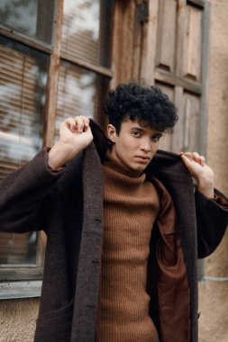 Kıvırcık saçlı, kahverengi paltolu ve kaplumbağa boyunlu genç adam eski tuğla binanın önünde duruyor. moda, tarz konsepti.