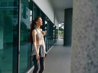 Spor kıyafetli, uzun saçlı genç bir kadın modern cam mimarisinin dışında bir anlık mutluluktan zevk alıyor.