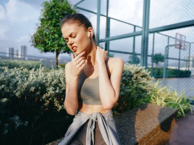 A woman in a sports bra and leggings touches her neck, showing discomfort against a backdrop of greenery and a basketball court clipart