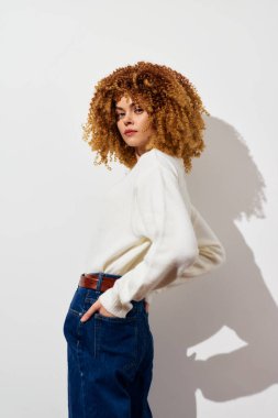 Şehvetli kıvırcık saçlı, yumuşak beyaz bir kazak ve sıradan bir kot pantolon giymiş, kendine güvenen, çağdaş moda estetiğini somutlaştıran, kendine güvenen genç bir kadın.