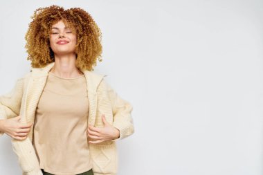Kendine güvenen genç bir kadın şık bej süveterini, kıvırcık saçlarını ve neşeli ifadesini, yaratıcı görseller ve moda tanıtımı için ideal, tarafsız bir arka plana karşı sergiliyor.