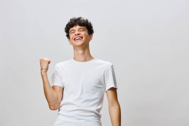 Sade beyaz bir tişört giyerken heyecan ve başarı ifade eden kıvırcık saçlı neşeli bir genç adam açık gri arka plan onun neşeli tavırlarını vurguluyor ve hoş bir atmosfer yaratıyor.