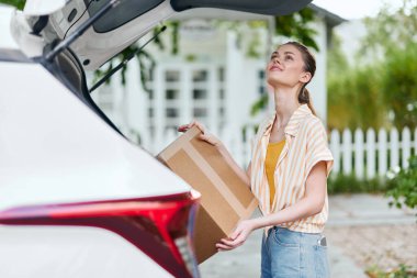 Genç bir kadın bir arabanın açık bagajında duruyor, elinde gülümsemeli bir paket var. Güzel bir açık hava ortamı, teslimat temaları, heyecan ve çağdaş yaşam tarzı sunuyor.