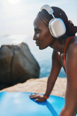 Örgülü saçlı, büyük gümüş kulaklıklı, kollarını mavi yoga minderine koyan bir kadının profili. Etrafı kayalık araziyle çevrili ve arka planda geniş bir okyanus manzarası var.