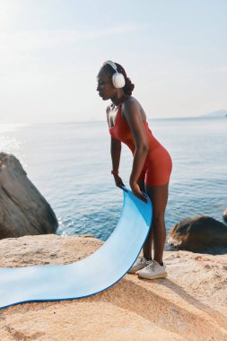 Aktif kıyafetli, kulaklıklı genç bir kadın sakin okyanusun kıyısındaki kayalık bir kıyıda mavi yoga minderi seriyor.
