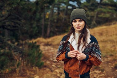 Şık şapkalı ve ceketli genç bir kadın huzurlu bir ormanda, toprak bir yolda zarif bir şekilde duruyor.