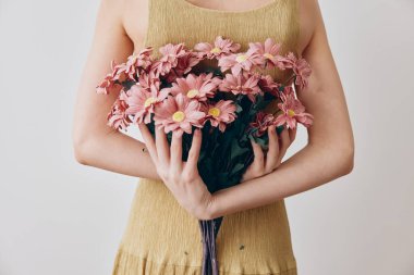 Beyaz arka planda pembe papatyalar tutan sarı elbiseli kadın. Parlak kıyafetli zarif bayanın ellerinde güzel bahar çiçekleri. Tasarım projeleri için büyüleyici ve taze görüntü.