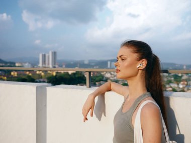 Uzun saçlı, kolsuz bluz giyen bir kadın korkuluklara yaslanmış, manzaralı bir şehir zeminiyle rahatlamış görünüyor.