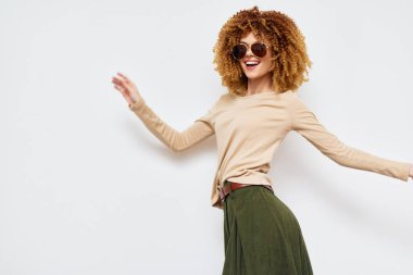 Açık bej bir bluz ve yeşil bir etek giyen neşeli bir kadın, kendine güven ve neşe saçan şık bir güneş gözlüğü takıyor ve beyaz bir arka plana şakayla poz veriyor.