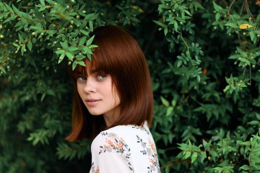 Canlı kızıl saçlı kadın yapraklarıyla yemyeşil çalıların önünde zarif bir şekilde duruyor.