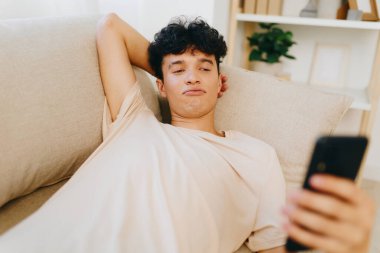 Bej bir kanepede uzanan, akıllı telefonuyla meşgul olan genç bir adamın gündelik ortamında düşünceli bir yüz ifadesi sergiliyor, evde dinlenme ve teknoloji kullanımı ile ilgili bir anı aktarıyor.