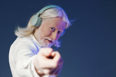 Uzun beyaz saçlı neşeli yaşlı bir adam, kulaklık takıyor, izleyiciye şakacı bir şekilde işaret ediyor. Görüntü müziğin hareketli ve ilgi çekici atmosferini arttıran sağlam bir mavi arkaplan içeriyor.