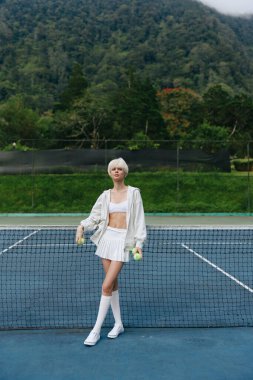 Tenis kortunda şık bir beyaz kadın modeli, kendine güven ve yaz enerjisi yayıyor, yemyeşil bir geçmişi ve canlı bir atmosferi var.