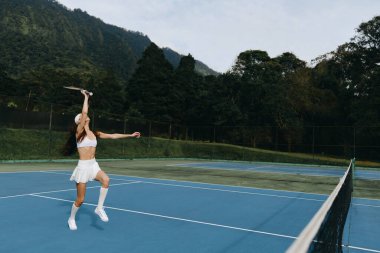 Mavi tenis kortunda görev yapan, beyaz etekli ve üstsüz bir kadının dinamik sahnesi arka plandaki yemyeşil dağlar aktif bir yaşam tarzını ve spor sağlığını vurguluyor.