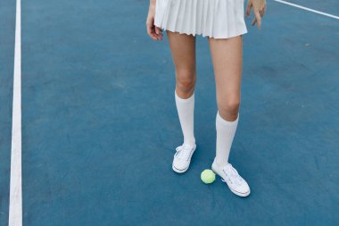 Beyaz etek ve diz boyu çorap giymiş genç bir bayan tenisçi, mavi tenis kortunda sarı bir tenis topunun yanında hazır, özgüven ve enerji saçıyor.