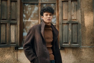 Kahverengi ceketli ve balıkçı yaka kazaklı şık bir genç, pencereleri kapalı, eski bir binanın önünde dikiliyor..