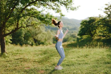 Şık giyimli genç bir kadın yemyeşil bir parkta neşeli bir şekilde ip atlıyor. Doğadaki sağlıklı yaşam tarzını ve uygunluğunu vurguluyor.