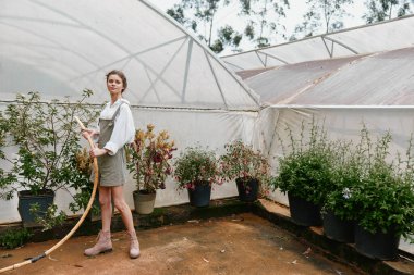 Günlük kıyafetli genç bir kadın yemyeşil bir serada neşe içinde bitkileri suluyor, doğa ve sürdürülebilirlik sevgisi somutlaştırıyor çevre dostu ve bahçıvanlık kavramları için ideal.