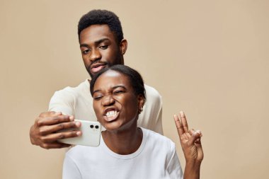 Mutlu genç bir siyah çift, bej bir arka plana karşı neşeli ifadelerle selfie çekmekten zevk alır. Neşeli ruh halleri, aşkı, bağlantıyı ve modern ilişkileri simgeler.