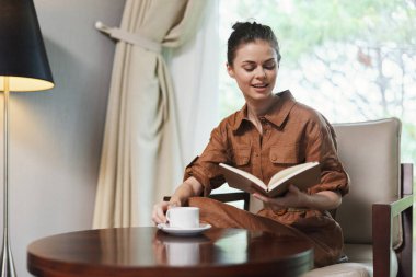 Bu resim, rahat kahverengi bir tulum giymiş, şık bir sandalyede oturmuş, kitap okuyup, kahve yudumlarken, huzur ve rahatlık içinde,