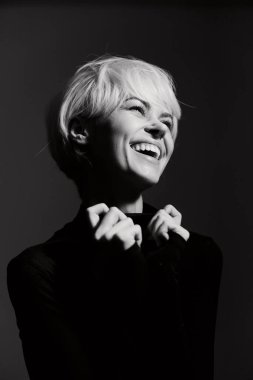 Kısa sarı saçlı, gülümseyen kadın siyah bir bluz giyerken mutluluk ve kendine güven gösteriyor. Koyu bir arkaplan üzerine kurulmuş bu resim neşe ve pozitiflik taşır.